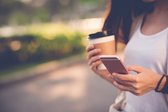 A woman walking in a park holding a cup of coffee and texting on a smart phone.